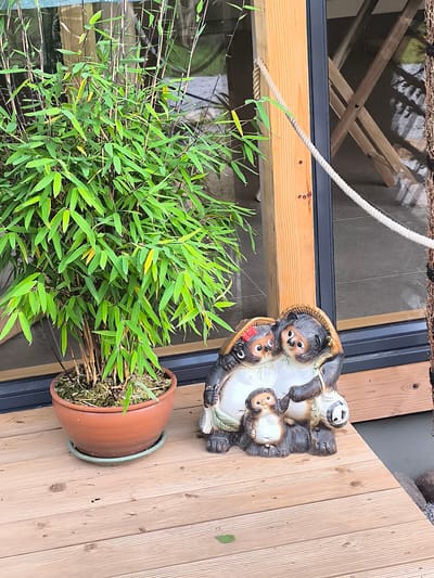 This image depicts an outdoor scene on a wooden deck, likely in Mažučiai, Lithuania. A potted bamboo plant with lush green foliage stands in a terracotta pot on the left. To the right of the plant are three ceramic raccoon dog (tanuki) figurines arranged in a group. The largest two figurines are positioned behind a smaller one, with the largest on the right holding a sake flask. The figurines have exaggerated features characteristic of tanuki statues, including wide smiles and round bellies. A thick, light-colored rope is visible extending diagonally across the image, attached to a wooden beam on the right. In the background, a building with large windows and wooden architectural elements can be seen. The overall setting suggests an outdoor seating area or entrance to a establishment.