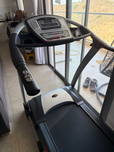The image shows a treadmill in a residential setting, possibly a living room or home gym, with a large glass sliding door in the background that opens to an outdoor balcony or patio. The balcony overlooks a dry, hilly landscape with sparse vegetation, indicative of an arid or Mediterranean climate. Through the glass, a portion of the city of Sderot, Israel, can be observed.
The treadmill is a York Fitness model, silver and black in color, with a digital display panel showing various workout metrics. It has a handrail with speed and incline adjustment buttons. In the foreground, a portion of a grey sofa is visible. On the tiled floor outside the glass door, a pair of grey athletic shoes with red laces and grey socks are placed together. A yellow electrical cord is visible on the floor, and part of a hanging chair structure is also present on the patio. The interior space appears to be a tiled floor.