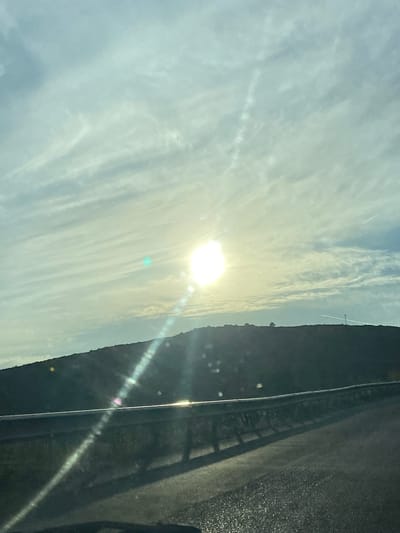 This is a horizontal, outdoor, daytime, eye-level shot of a sunlit landscape with a road in the foreground. The sun is bright and positioned slightly left of center in the upper portion of the frame, casting lens flares and a bright glare across the sky and down onto the road. The sky is filled with wispy, streaky clouds that are tinged with yellow and blue hues from the sunlight.

In the midground, a dark, silhouetted hillside stretches across the frame. A thin, white streak, possibly a contrail from an aircraft, is visible against the sky above the right side of the hillside. Two small, dark shapes, one round and one elongated, are visible on the crest of the hill, possibly structures or trees.

The foreground consists of a dark asphalt road curving to the right. A metal guardrail runs along the edge of the road, catching the sunlight and reflecting a bright glare. The road surface is textured and appears to be damp or slightly wet in places, reflecting the light. The overall impression is of a scenic drive, possibly on a clear but overcast day with the sun breaking through the clouds. The specific location in the city of Višnjevo, Montenegro, cannot be determined from the visual information alone.