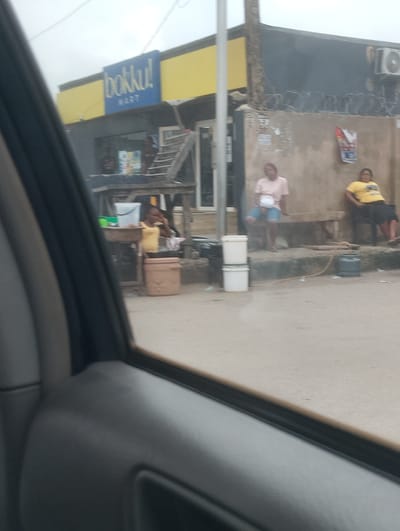 The image shows a street scene in Alagbede, Nigeria. In the foreground, the interior of a vehicle is visible, with a dark gray door panel and part of a window. Outside the vehicle, a building with a yellow facade and a blue sign reading "bokku! MART" is visible. Below the sign, a dark-colored structure with wooden elements is present. In front of this structure, a person in a yellow top and dark skirt is seated on a bench, and another person in a light pink top and blue shorts is also seated on a bench. Several white buckets are stacked on the ground, and a gray gas cylinder is also visible. A concrete wall with barbed wire runs along the back of the scene, and posters are attached to it. The ground is a sandy or dirt surface. The sky is overcast.