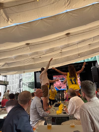 The image depicts an outdoor gathering under a white canopy, likely a beer garden or event space. In the background, a large screen displays a sports broadcast, featuring close-ups of male athletes. The foreground shows several people seated at tables, some with drinks, observing the screen. Two individuals are standing on raised platforms in front of the screen, one in a yellow shirt with the name "BROLIN" and the number "11" visible, gesturing towards the audience. The other individual, wearing a light-colored shirt and brown shorts, is also standing on a platform. The overall atmosphere appears festive, with a focus on the sports event. The presence of "FOYNHA GEN" on a banner suggests a location within or associated with this establishment. Tønsberg, Norway, is mentioned as the city context.