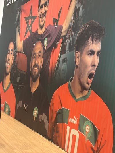 The image is a photograph of a poster featuring three Moroccan soccer players. The player at the top is wearing an orange Puma jersey with the number 10 and the Moroccan soccer federation logo. He is yelling with his mouth open. Below him, two players are holding a Moroccan flag, both wearing purple jerseys with the Moroccan soccer federation logo. The background is blurred, suggesting a large crowd. The image does not contain enough information to describe Ouahat Sidi Brahim, Morocco.
