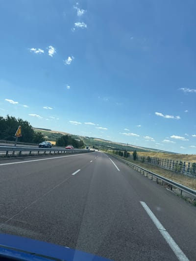 The image shows a view from a vehicle traveling on a highway in the rural area surrounding Bouleuse, France. The sky is bright blue with scattered white clouds. The highway has asphalt pavement and is divided by white lane markings. Several cars are visible on the road. On the left side of the highway, there is a guardrail, trees, and fields. On the right side, there is a metal fence, dry grass, and trees in the distance. A yellow warning sign with a depiction of a person crossing the road is visible on a pole on the left side of the highway. The landscape is hilly and appears to be agricultural or natural terrain.
