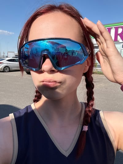 A woman is standing outside on a sunny day, with a light-colored sky visible in the background. She has auburn hair styled in braids. She is wearing large, reflective blue sunglasses that obscure her eyes. Her left hand is raised towards her forehead. She is wearing a dark blue top with light-colored trim. Cars are parked in the background. The location is Казань, Russia, as identified by a sign with Cyrillic characters. The setting appears to be a parking lot or street near a commercial building.