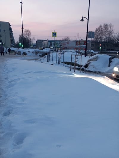 The image presents a snow-covered urban landscape in Yaromaska, Russia, observed during twilight hours. The foreground is dominated by undisturbed snow, forming what appears to be a pedestrian path, with isolated footprints visible. To the immediate left, a cleared section of the path shows four individuals, dressed in dark winter clothing, walking away from the camera. In the background on the left, a multi-story residential building with a light-colored facade is visible. A tall street lamp stands near this building, and a traffic light displays a green signal.

Towards the center and right, a series of young, bare trees, each supported by wooden stakes, line a snow-covered median. Large, irregular mounds of snow, likely accumulated from plowing, are present. A black, ornate street lamp extends over the street on the right, illuminating a portion of the snow. A white vehicle, with its headlights illuminated, is partially visible in the bottom right corner, indicating its presence on the roadway. Directly across the street, a yellow pedestrian crossing sign is prominent. Behind it, a commercial building features a red and white facade with the Cyrillic text "МЕБЕЛЬ" (Furniture) clearly visible. Further down the street on the right, another traffic light displays a red signal. Deciduous trees, devoid of leaves, are scattered throughout the background. The sky exhibits a pale pink and light blue gradient, characteristic of dawn or dusk.