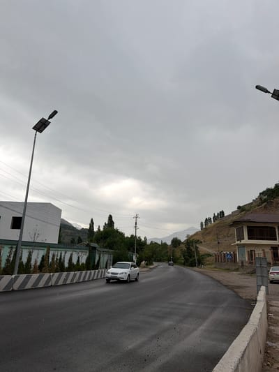 The image depicts a street scene in the city of Khumsan, Uzbekistan, under an overcast sky. The road is asphalt, wet from recent rain, with a concrete barrier along the right side. On the left, a white sedan is driving away from the camera. Beyond the road, a white building with a dark window opening is visible, bordered by a fence and rows of green shrubs. A tall lamp post with a solar panel stands to the left of the building.

Further down the road, a second car is visible in the distance. To the right of the road, a brown-roofed building with large windows is situated on a steep, grassy hillside dotted with trees. A pedestrian crossing sign is visible near this building. The landscape rises into mountains in the background, partly obscured by clouds. Power lines and utility poles are present throughout the scene. The overall impression is of a somewhat quiet, possibly rural or suburban, area with infrastructure for street lighting and residential or commercial buildings.