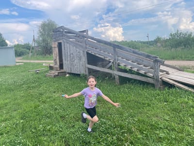A young girl with brown hair, wearing a purple t-shirt and black shorts, runs across a grassy field. She has a joyful expression and is holding a blue object in her left hand and a clear plastic cup in her right hand. Behind her is a large wooden structure with a ramp, and in the background, there are trees and a cloudy sky. The scene appears to be in a rural setting in Nizhnii Bugrysh, Russia.