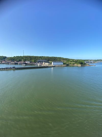 The image depicts a waterfront scene in Gothenburg, Sweden, on a clear, sunny day, likely in the late morning or early afternoon given the bright sunlight and distinct shadows. The perspective is from a high vantage point, looking down and across a body of water.

The main subjects are the canal or river and the adjacent shoreline. On the land side, there is a dense green forest bordering the water, with a built-up area interspersed within the trees. This area appears to be a small town or port, featuring various buildings, some with red roofs, and docks with several boats moored. A prominent large, white, rectangular building with many windows stands out near the water's edge. Further along the coastline, more structures and infrastructure, including what looks like a tall transmission tower, are visible. The water itself is a greenish-brown color, with visible ripples indicating a slight breeze.

The overall activity is that of a functioning port or waterway, with boats at anchor and buildings suggesting human habitation and industrial or commercial activity. The clear blue sky and bright sunlight indicate fair weather. The location cues, such as the style of architecture and the general landscape, are consistent with a Scandinavian coastal town. No specific emotions are conveyed by the scene, which is presented in a factual, observational manner. There is no discernible text in the image.