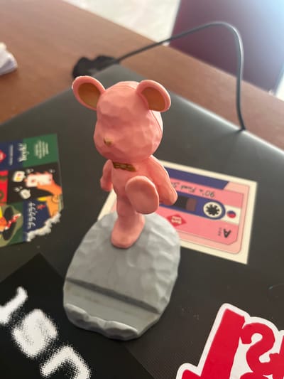 A pink figurine of a bear is lying on its back on a black surface, possibly a laptop. The bear is positioned on top of a gray, rock-like object. To the left of the bear is a sticker of a pink cassette tape with the text "90's Funk" and "A side" visible. To the right of the bear, partially visible, is a sticker with large red lettering. Below the cassette tape sticker, a collage of smaller stickers is visible, featuring various cartoonish images and text. A black cable extends from the upper left of the frame. The overall scene appears to be a casual arrangement on a desk, illuminated by overhead lighting, suggesting it is daytime.