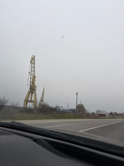 The image depicts an overcast, grey day in 2 Mai, Romania, viewed from the interior of a vehicle looking forward through the windshield. The dark dashboard and the base of the windshield are visible in the lower foreground. An asphalt road, defined by a solid white line on its right edge, occupies the mid-ground. To the left of the road, a strip of sparse brown grass leads to a grey, rectangular concrete fence. Behind this fence, two prominent industrial gantry cranes, colored yellow, stand tall; the foremost crane appears taller and more defined, featuring cables and a hook mechanism. Leafless bushes and small trees are interspersed along the fence line and around the cranes. Further right, a tall, dark-colored metallic communication tower with multiple antenna arrays and a ladder system is visible. Beyond the tower and to the far right, several low-rise structures, likely industrial or residential, with white walls and red roofs, are present. The sky is uniformly light grey, indicating an overcast condition. No individuals, explicit actions, or direct interactions are visible within the scene.