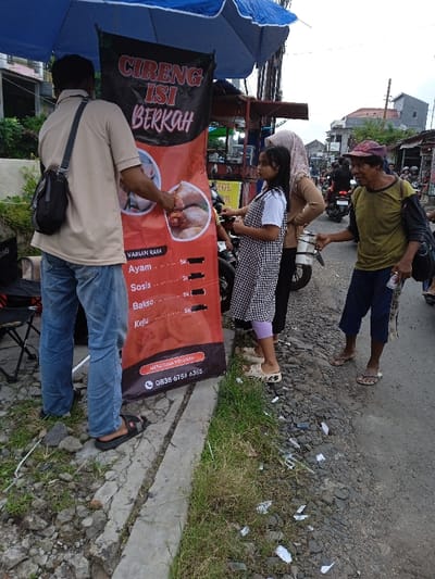 The image depicts a street scene in the city of Kauman, Indonesia, focusing on a roadside food stall and several individuals.
In the foreground, slightly to the left, a man stands with his back partially to the viewer, wearing a light brown t-shirt, blue jeans, and black flip-flops. A black shoulder bag with a gray strap is slung over his right shoulder. He is positioned in front of a red and black banner advertising "CIRENG ISI BERKAH" (Filled Cireng Blessing), which displays images of food, "VARIAN RASA" (Flavor Variations) including Ayam, Sosis, Bakso, and Keju, each priced at "5k," and a contact number for orders. The man appears to be touching or presenting something on the banner.
To the right of the banner, a young girl, approximately 8-12 years old, stands facing right. She wears a black and white checkered dress over a white top, light purple leggings, and white patterned sandals. Behind her, a woman in a light brown long-sleeved top and a light-colored hijab is partially visible. A white scooter or motorcycle is parked behind the woman.
Further to the right, an older man with a weathered appearance stands facing the girl and woman. He wears a mustard yellow t-shirt, dark blue cropped pants, a red cap, and brown flip-flops. He extends his right hand forward, holding a white plastic cup, while his left hand holds a crumpled white plastic bottle.
The environment includes a rough asphalt or gravel road surface with a cracked concrete curb, and patches of green grass and scattered white litter along the roadside. Overhead, a large blue umbrella provides shade for the stall area. In the background, beyond the immediate subjects, a busy street is visible with additional people and several motorcycles, along with residential or commercial buildings and overhead utility wires under an overcast sky.