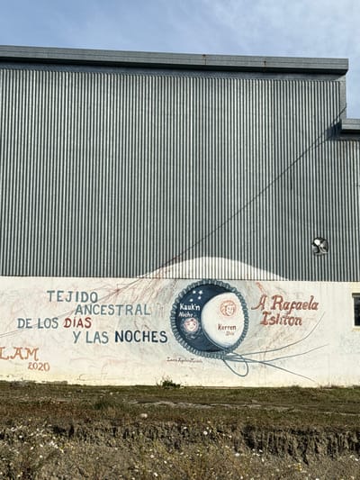 The image displays a section of a building located in Tolhuin, Argentina, beneath an overcast sky. The upper part of the structure consists of vertical corrugated metal siding. A dark utility cable extends diagonally from the upper left, crossing the corrugated metal, and descends into the lower painted section of the wall. A small, dark, circular fixture is mounted on the corrugated metal in the upper right portion.
The lower section of the building presents a white-painted wall with a mural. Prominent text on the left reads "TEJIDO ANCESTRAL DE LOS DÍAS Y LAS NOCHES". Below this, "LAM 2020" is visible. On the right, more decorative text states "A Rafaela Ishton". Below the central mural element, the artist's signature "Laura Aguilera Miranda" is present. The central motif is a large circular design with a gear-like border, horizontally bisected. The left half is dark blue with stars and a stylized face, labeled "Kauk'n Noche". The right half is lighter, displaying a different stylized face, labeled "Kerren Dia".
The foreground features an unmanicured area of dry earth with sparse, light-colored vegetation and scattered small white flowers.