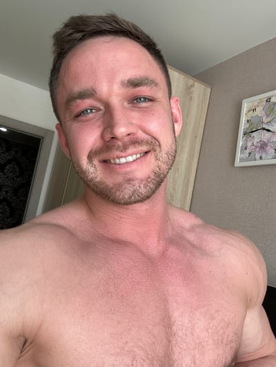 A shirtless man with short brown hair, blue eyes, and a trimmed beard is smiling at the camera. He appears to be indoors. A clock with a floral design hangs on the wall behind him. A doorway is visible in the background. The city of Dobryanka, Russia is not evident from the image.