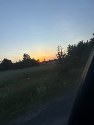The image depicts a rural scene at sunset, with the sun appearing as a small, bright orange orb low on the horizon. The sky transitions from a clear, pale blue at the top to hues of orange and yellow near the sunset. Silhouetted trees form a dark line across the middle ground, interspersed with a utility pole topped with a single crossbar. To the right of the trees, a grassy field stretches into the distance, appearing darker and less defined due to the fading light. A diagonal dark band, possibly from a window or a car interior, cuts across the upper right portion of the image, suggesting the photo was taken from inside a vehicle. The overall impression is one of a peaceful, natural landscape at the end of the day. The time of day is clearly sunset. The weather appears clear.