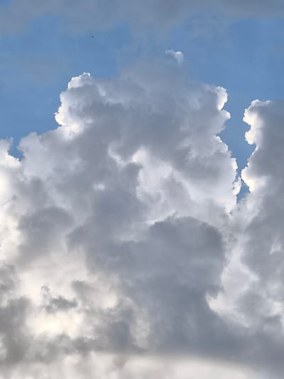 The image depicts a close-up view of dramatic, cumulus clouds against a bright blue sky. The clouds are illuminated by sunlight, creating strong contrasts between bright whites and dark grays, suggesting a dynamic weather system. Some clouds exhibit sharp, illuminated edges, while others are massed and appear dense. In the upper left quadrant, a tiny dark object, possibly a bird or an aircraft, is visible against the blue sky. The overall scene is natural and atmospheric, with no discernible urban elements or specific landmarks of Broek op Langedijk, Netherlands. The perspective is upward, focusing solely on the sky and cloud formations.