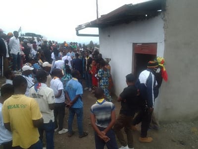 A large diverse crowd is gathered near a small, white building. Some individuals wear Scout uniforms with yellow or blue shirts. Others are in casual attire, including dresses and jeans. A vehicle is visible in the background. A man wearing a mask and a hat with red, yellow, and black colors stands near a doorway. The scene appears to be outdoors, potentially on a hillside or elevated area.