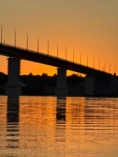 The image captures a bridge silhouetted against a vibrant orange sunset over the Kama River in Kambarka, Russia. The bridge, a significant piece of infrastructure, features a series of lampposts marching along its length, casting long shadows on the water below. The river's surface is textured with ripples, reflecting the warm hues of the sky and the dark shapes of the bridge's supports and the distant tree line. The atmosphere is tranquil and serene, characteristic of a peaceful evening in this riverside city. There are no people visible in the frame.