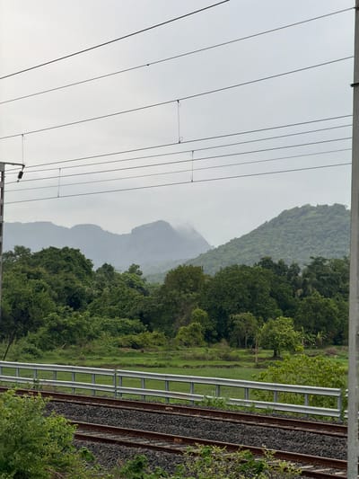The image captures a scenic view of mountains shrouded in mist, with lush green vegetation in the foreground. Train tracks run horizontally across the lower portion of the frame, bordered by a metal guardrail and a grassy field. Overhead, a network of electrical wires stretches across the sky, suggesting the presence of a railway line. The overall atmosphere is serene, indicative of a cloudy or rainy day, possibly in the vicinity of Vasai-Virar, India, given the terrain and infrastructure. There are no people or animals visible in the image. The focus is on the natural landscape and the man-made railway system.