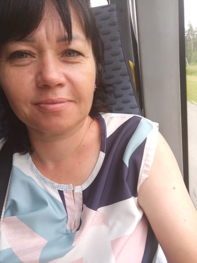 A middle-aged Caucasian woman with dark brown hair and green eyes is seated on what appears to be a bus, judging by the textured seat behind her and the window showing a rural landscape. She is looking directly at the camera, and her expression is neutral. She is wearing a brightly patterned shirt with geometric shapes in shades of white, navy, light blue, and pink. A small pink earring is visible in her right ear. The environment outside the window suggests a natural setting with trees and a road. The lighting indicates it is daytime.