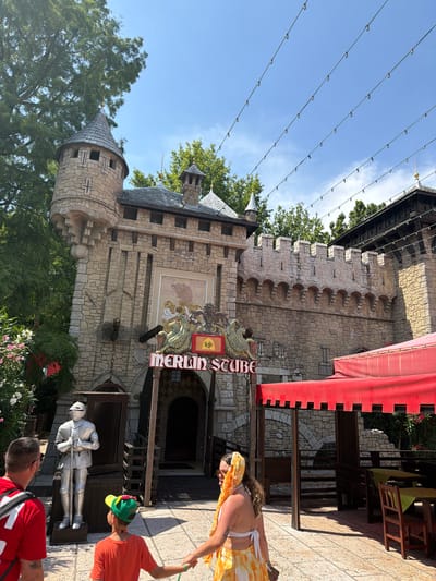 The image shows the exterior of a medieval-themed building, likely a restaurant or attraction, with a castle facade and a sign that reads "MERLIN STUBE". The building is made of stone with turrets and battlements. A statue of a knight in armor stands to the left of the entrance. In the foreground, two people are visible: a man in a red shirt and a child in an orange shirt and a green hat. A woman in a yellow and white outfit is walking past them. The scene is outdoors with trees and a bright blue sky. String lights are visible overhead. The location is identified as Castelnuovo del Garda, Italy.