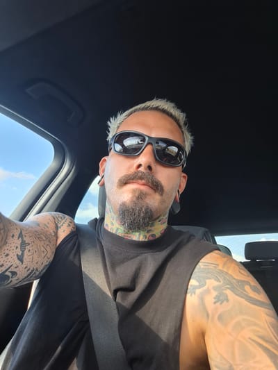 An individual with blonde, spiky hair and a goatee is sitting in the driver's seat of a car. They are wearing sunglasses, a black t-shirt, and a seatbelt. The individual has numerous tattoos on their arms and neck, including what appears to be a lizard tattoo on their right arm. The car interior is dark, with the sky and clouds visible through the windows. The image appears to have been taken during the daytime, likely on a sunny day, suggesting an outdoor location. The context of Shtarkovo, Bulgaria, is not discernible from the image itself.