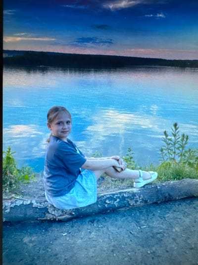 A young girl is sitting on a fallen log near the edge of a body of water. The girl has blonde hair pulled back in a ponytail and is wearing a grey t-shirt and a light blue skirt. She is looking towards the camera with her arms resting on her knees. The water is calm and reflects the blue sky and some clouds. The shoreline is lined with trees and greenery. The sky transitions from a deep blue at the top to lighter shades of pink and orange near the horizon. The image appears to be taken during dusk or dawn. The girl's pose and the serene environment suggest a moment of quiet reflection. The location is not explicitly stated in the image, and there is no information provided about Protasovo, Russia.