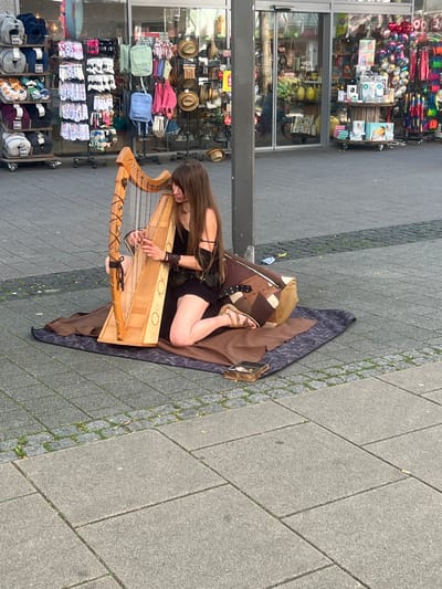 A woman is playing a harp on a sidewalk in front of a store in Herne, Germany. She is kneeling on a blanket with a large drum next to her. The store behind her sells various items, including clothing, hats, and bags. The sidewalk is paved with cobblestones and there are some patches of grass. The woman has long brown hair and is wearing a black dress. She is focused on playing the harp and her hands are on the strings. The drum is covered in a patterned fabric and has some buckles and straps. The store has large glass windows and a sign above the entrance. The image is taken from a slightly elevated angle, looking down at the scene.