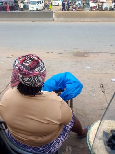 A woman is seated on a chair on a concrete surface next to a road in Ihiala, Nigeria. She has a colorful patterned headscarf, a tan top, and patterned leggings. A blue plastic bag is next to her. The road has a median and vehicles are visible in the background. Other people and stalls can be seen on the other side of the road. A woven basket is on the ground near the woman.