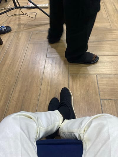 The image shows two people sitting on a wooden floor. One person is wearing black pants and black sandals, while the other is wearing white pants and black shoes with white soles. A dark blue object is visible in the lap of the person wearing white pants. There is also a camera light setup on the left side of the frame.