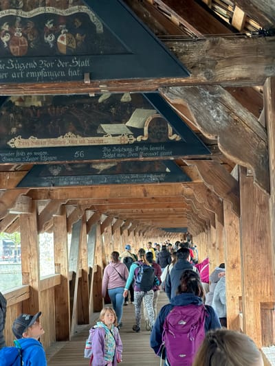 The image captures the interior of the Chapel Bridge in Lucerne, Switzerland. The bridge is characterized by its historic wooden structure, and it displays a series of triangular paintings hung below the roof. Several people are walking along the bridge's wooden walkway, including a girl looking towards the camera and a boy with a baseball cap looking upwards, apparently at the paintings. The paintings feature German text and heraldic symbols.