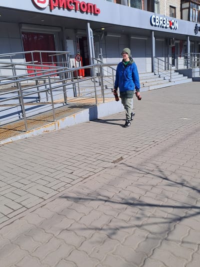 In Меткомелино, Russia, an outdoor scene depicts a single individual walking on a paved sidewalk in front of two commercial storefronts. The individual, appearing male, wears a dark green beanie, glasses, a blue puffer jacket over a green hooded sweatshirt, light grey trousers, and black Adidas sneakers. They carry one red bottle with a black cap in each hand.
To the left of the individual is a storefront with the red and white "БРИСТОЛЬ" sign, featuring a closed red roller shutter and an accessible ramp constructed of corrugated metal with stainless steel handrails, showing signs of rust. A red cylindrical object with a white 'Б' symbol is positioned near the entrance. To the right, another storefront bears the blue and white "СВЯЗЬОN" sign, with multiple windows and doors obscured by what appear to be closed roller shutters, accessed by concrete steps also fitted with stainless steel handrails. An air conditioning unit is mounted above this storefront.
The sidewalk is paved with grey interlocking concrete blocks, transitioning from rectangular patterns closer to the buildings to a wavier pattern further out. Long, indistinct shadows are cast across the sidewalk from the right, suggesting bright sunlight. The sky is clear and light blue.