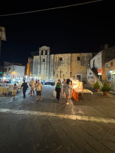 The image depicts an outdoor market scene at night in the city of Orosei, Italy. In the background, a weathered, ancient building, likely a church or historical structure, dominates the scene with its stone facade and bell tower. Several people are walking through the cobblestone square where market stalls are set up. One stall on the right displays various items, possibly souvenirs or crafts, illuminated by artificial lighting. People are seen browsing the stalls and walking around, engaged in evening activities. The atmosphere suggests a lively town square during a nighttime market.