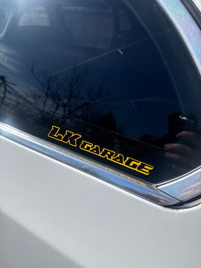 This image depicts a close-up of the rear driver's side window and adjacent body panel of a light grey or silver vehicle, observed in Daskal-Atanasovo, Bulgaria. Affixed to the lower portion of the dark-tinted window is a yellow adhesive decal spelling "LK GARAGE" in capitalized block letters with a black outline. The window surface exhibits multiple minor scratches, dust, and streaking. Reflected in the glass are bare tree branches against a bright, clear sky, indicating an outdoor, daytime setting. A partial reflection of a hand holding a dark-colored smartphone, likely the device capturing the image, is visible in the lower right corner of the window. A chrome or silver trim piece separates the window from the vehicle's body panel, which appears clean and unblemished within the frame. No individuals are directly visible.