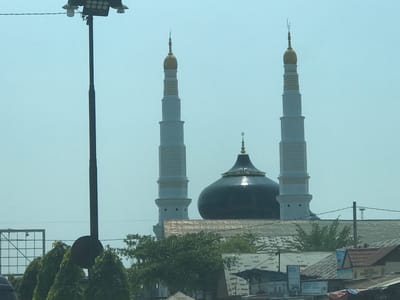The image presents a view of the Baiturrahman Grand Mosque located in Keudematangglumpangdua, Indonesia. Two white minarets with gold finials flank a dark-colored central dome, which is surmounted by a crescent moon symbol. The mosque is partially obscured by trees and the roofs of buildings, giving the impression of being integrated into the urban landscape. A tall, dark-colored utility pole with multiple lights is visible in the foreground, obstructing some of the view. The overall environment suggests a daytime scene with a slightly hazy atmosphere.