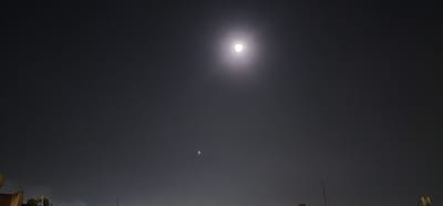 The image is a night photograph taken in Sderot, Israel, featuring a dark sky illuminated by a bright full moon. The moon is positioned in the upper center of the frame, emitting a soft glow that creates a hazy halo around it. Several faint stars are scattered across the sky. In the lower portion of the image, the silhouettes of buildings and satellite dishes are visible, suggesting an urban or suburban setting. One of the satellite dishes, located on the left side, is particularly prominent, with a portion of a building's facade beneath it. To the right, a thin vertical pole rises from the darkness. A small, indistinct circular object is visible in the sky, slightly below and to the right of the moon. The overall atmosphere is dark and serene, with the moonlight casting a dim, atmospheric light over the scene.