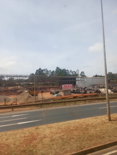 This image depicts an outdoor industrial or construction site located in Kikuyu Township ward, Kenya, viewed from across a multi-lane asphalt road under a partly cloudy sky.

In the immediate foreground, a reddish-brown dirt shoulder with sparse, dried vegetation separates the viewer's perspective from the paved road. The road features three visible white arrow markings indicating forward vehicular movement. A metal guardrail runs parallel to the road, forming a boundary with the industrial area. To the right, a tall, slender grey utility pole extends vertically out of the frame.

Beyond the road and guardrail, the primary subject is an active site. A prominent red and white sign displays "K PIPES HARDWARE." Adjacent to it, a small, light-colored structure with a dark roof is visible. The site contains numerous dark, cylindrical pipes stacked horizontally, along with several white cylindrical tanks or containers. Large piles of reddish-brown dirt and grey gravel are present. A yellow construction vehicle, possibly a loader or excavator, is parked near these piles, and a white truck is partially visible further left. In the background of the site, a large building framework under construction is partially covered with white sheeting, featuring extensive metal scaffolding or structural elements. Dense green trees form a natural backdrop behind the construction activity. No individuals, explicit actions, or interactions are discernible within the scene.