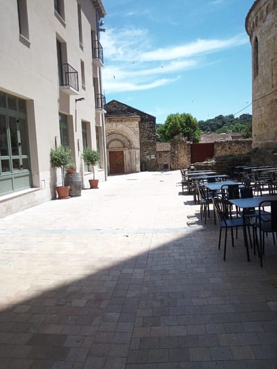 The image depicts an outdoor seating area in a courtyard with tiled flooring. To the left, a light-colored building with modern windows and balconies is visible. In the center, a stone archway leads to a building with a wooden door, and two potted plants flank the entrance. To the right, a round stone structure with arched windows stands, partially obscuring a view of a treelined hillside. Numerous tables and chairs, set up for outdoor dining, occupy the right side of the courtyard. The sky is blue with scattered white clouds. The overall scene suggests a commercial establishment, possibly a restaurant or cafe, in a historic urban setting. The architecture and paving are consistent with the medieval town of Besalú, Spain, known for its well-preserved Romanesque and Gothic structures, particularly its iconic bridge.