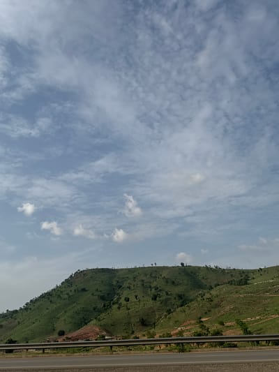 The image captures a landscape near Kugbo, Nigeria. A grassy, tree-dotted hillside dominates the right side of the frame, transitioning into a road with multiple lanes. To the left, the sky is filled with a mix of light blue and white clouds. The perspective is slightly tilted. There are no people visible. The scene appears to be outdoors during daylight hours.