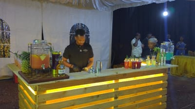 An event scene displays a bartender standing behind a custom-built bar with a wooden pallet design and integrated lighting. The bar is lined with various drinks and cocktail ingredients, including juice dispensers, liquor bottles, and garnished cocktails in plastic cups. Several people stand in the background, suggesting a social gathering or celebration. The setting appears to be indoors with white fabric draped overhead and dark curtains behind.