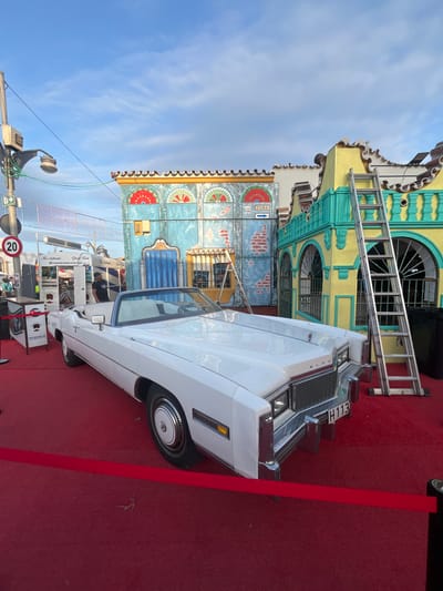 This image captures a vibrant outdoor scene, likely a festival or themed event, in Fuengirola, Spain. The primary subject is a pristine white, classic American convertible, possibly a Cadillac Eldorado ("ELDO" visible on the grille), parked on a bright red carpet. Its license plate reads "H113".
In the background, an elaborate, multi-story building facade dominates the scene with its vivid colors of yellow, turquoise, and blue, featuring intricate architectural details, arched windows, and decorative flourishes. A silver ladder leans against the building. Further left on the building, a sign with "CUBA" and an arrow is visible, suggesting a Cuban or Latin American theme for the setting.
To the left of the car, partially obscured, is a street food stand with signage that reads "The Authentic Street Food" and "la pequeña habana street food," along with a phone number "Telf: 605 899 421". A few people can be seen near this stand, and one person appears to be working. A red circular traffic sign indicating "20" (likely a speed limit) is also visible on the far left.
The sky is a clear blue with wispy clouds, indicating good daytime weather. The red carpet extends across the ground, enhancing the festive or grand event atmosphere. The overall impression is one of celebration, with a blend of classic automotive culture and themed street fair elements.