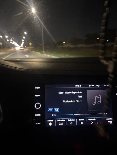 The image depicts a nighttime scene from the interior of a vehicle located in Komchén, Mexico. The view is forward through a dirty windshield, revealing a multi-lane paved road illuminated by numerous streetlights extending into the distance on the left. The headlights of at least one distant vehicle are visible. A yellow, diamond-shaped road sign is partially discernible on the right side of the road, with vegetation bordering the roadside. The vehicle's dashboard, a dark, textured surface, occupies the lower foreground. A prominent infotainment screen displays the time as 19:54, an ambient temperature of 25.5°C, and is actively playing the song "Noviembre Sin Ti" by Reik, with "Video disponible" indicated. The playback shows 1:15 elapsed with 2:08 remaining. Menu options such as RADIO, MEDIA, PHONE, VOICE, Fuente (with Bluetooth icon), Selección, and Ajustes are present on and around the screen. A dark, out-of-focus object, possibly a beaded hanging item, is visible on the far right, suspended near the windshield. No individuals are directly visible.