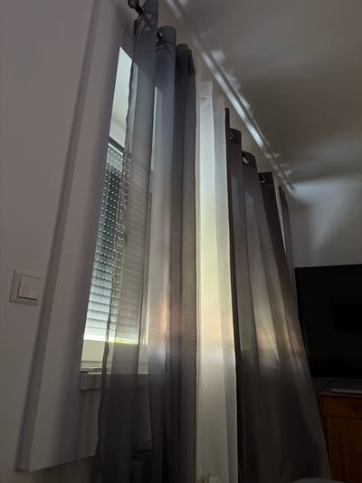 The image shows a room with light filtering through sheer curtains in shades of gray and white. The curtains are hung on a curtain rod above a window. The window has a slatted blind behind the curtains. A light switch is visible on a white wall to the left of the window. To the right, a dark television screen and a wooden cabinet are partially visible. Light patterns are cast on the ceiling. The scene appears to be an interior residential space. The location is identified as Weinheim, Germany, however, there are no visual elements in the image to corroborate this specific location.