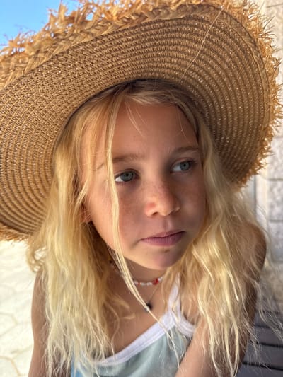 A young girl with blonde hair and freckles wears a straw hat and a necklace. She is looking to the right, with her face partially obscured by her hair and the hat. The background is blurred, showing what appears to be a light-colored, textured wall and a light blue sky. The image was taken outdoors, possibly in Kfar Vradim, Israel.