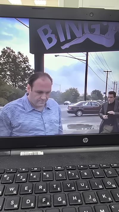 The image is a photograph taken from a laptop screen, showing a scene from a television show or movie. In the foreground, a man in a light blue collared shirt is looking down. To the right, another man in a dark jacket and light-colored pants is smoking a cigarette. Behind them, cars are visible on a street, and a sign with the word "BINGO" and a silhouette of a woman is displayed. The scene appears to be outdoors in a suburban or urban environment. The laptop's keyboard is visible in the lower portion of the image. There is no indication of the location being Oral, Kazakhstan.