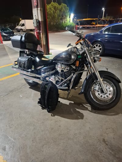 In Barri De Porta, Spain, a parked motorcycle with saddlebags and a top box sits on a concrete surface at night. The motorcycle is dark gray with chrome accents. A black backpack rests on the ground next to the rear wheel. In the background, a white van is parked, along with a dark-colored car. Further back, several yellow buses are lined up. The scene appears to be in a semi-industrial or service area, possibly a parking lot or a roadside stop, indicated by the presence of a fire extinguisher mounted on a pole and overhead lighting. The lighting suggests it is nighttime.
