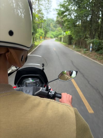 An over-the-shoulder shot shows a person riding a red scooter down a paved road lined with lush green trees and foliage. The rider wears a light-colored helmet and a tan-colored jacket. The scooter's dashboard, featuring a digital display with a green light, is visible. The road has a yellow line marking the center. In the distance, the road curves to the left, disappearing into the trees. A white and blue post stands on the right side of the road. The scene appears to be in a rural or semi-rural area, potentially Mae Hia Town Municipality, Thailand, given the dense vegetation and style of the road markers.