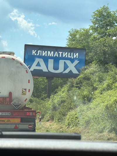 The image shows a billboard with the Cyrillic word "КЛИМАТИЦИ" ( Klimatitsi, meaning "air conditioners") above the brand name "AUX" in large white letters on a blue background. The billboard is situated in a rural or roadside environment, with lush green trees and foliage visible behind and around it. A white tanker truck with red accents and hazard symbols is parked to the left of the billboard, partially obstructing the view. The truck has license plates that appear to read "2018 EM". The sky is cloudy with patches of blue. The location is identified as Stefanovo, Bulgaria.