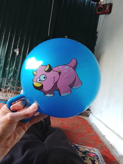 A person's hand is holding the blue handle of a blue inflatable ball with a cartoon pink dinosaur printed on it. The ball is large and round, with a small handle attached to the side. The dinosaur is pink with yellow horns and blue spots. The background is out of focus, but appears to be an outdoor setting. There is a corrugated metal fence on the left and a white wall on the right. A red rug is visible on the ground. The overall scene suggests a casual, perhaps playful, moment. It is impossible to determine the exact location within Mehnatabad, Tajikistan from the provided image.