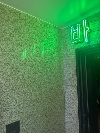 The scene depicts a corner entrance of a commercial establishment within Jongno-gu, South Korea. The environment consists of textured, light-grey speckled granite-like walls, illuminated by a dominant green neon light source. A bright green neon sign displaying the Korean character "바" (Ba), indicative of a "Bar" establishment, is affixed to the upper right wall, positioned above a dark-framed doorway. Faint, distorted green reflections of the neon characters are visible on the adjacent speckled wall to the left. A small, dark-colored square panel, possibly an intercom or access control device, with a faint green indicator light, is mounted low on the left wall. No individuals are present, and no actions or interactions are observed. The ceiling above is plain and white, reflecting the green light.
