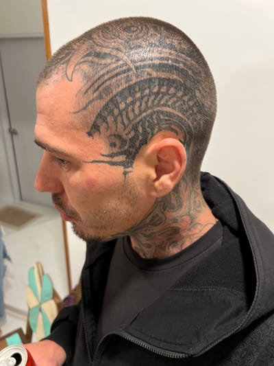 A man with close-cropped hair and extensive tattoos on his head, neck, and face is shown in profile. A large, swirling tribal-style tattoo dominates the side of his head. He wears a black hooded sweatshirt over a dark t-shirt. His facial hair is sparse and light in color. The background includes a white surface and a glimpse of other items, including a can. The image is rotated so the subject is lying on his side.